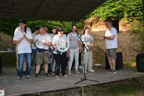 Pokaż zdjęcie: Grupa mężczyzn stojących na scenie. Na głowie mają czarne chusty z namalowanym kościotrupem. Dwóch z nich trzyma w ręku ster i małe koło ratunkowe. Śpiewają. 