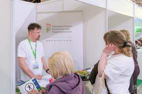 Pokaż zdjęcie: Pracownik stoiska PFRON udziela informacji grupie odwiedzających. Na ladzie znajdują się ulotki i materiały promocyjne.