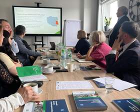Spotkanie przy stole konferencyjnym w ROPS w Lublinie. Uczestnicy słuchają prelekcji prezentowanej na ekranie o programach i dofinansowaniach PFRON; na stole materiały informacyjne, notesy, woda.