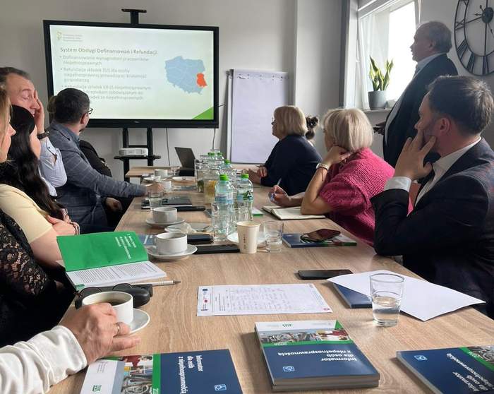 Pokaż zdjęcie: Spotkanie przy stole konferencyjnym w ROPS w Lublinie. Uczestnicy słuchają prelekcji prezentowanej na ekranie o programach i dofinansowaniach PFRON; na stole materiały informacyjne, notesy, woda.
