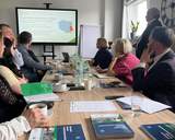 Spotkanie przy stole konferencyjnym w ROPS w Lublinie. Uczestnicy słuchają prelekcji prezentowanej na ekranie o programach i dofinansowaniach PFRON; na stole materiały informacyjne, notesy, woda.