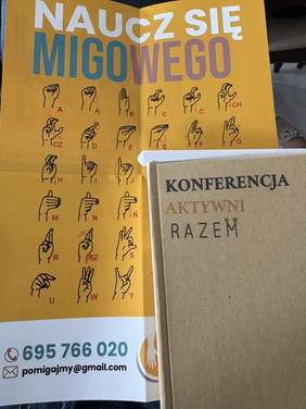 Pokaż zdjęcie: Na stole leży ulotka zachęcająca do nauki języka migowego. Po prawej stronie jest notes z napisem na okładce: Konferencja "Aktywni RAZEM". 