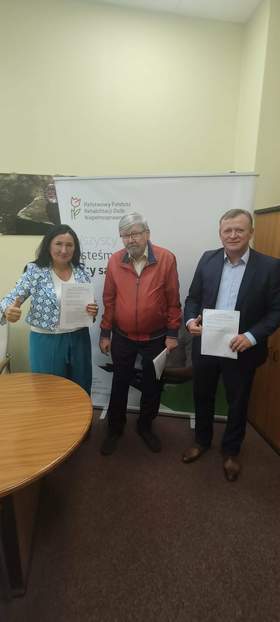 W gabinecie dyrektora Łódzkiego Oddziału PFRON stoi kobieta i dwóch mężczyzn. Zawarli umowy o dofinansowania. W tle widać baner z logo PFRON. 
