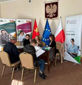 Pokaż zdjęcie: Trzy kobiety siedzą przy stole i rozmawiają z kobietą oraz mężczyzną naprzeciwko. W tle widoczne są banery Powiatowego Urzędu Pracy w Białymstoku i Państwowego Funduszu Rehabilitacji Osób Niepełnosprawnych oraz flagi Polski, Unii Europejskiej i Tykocina. Na stole znajdują się materiały informacyjne.