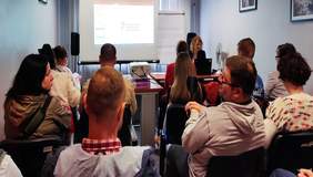 Pokaż zdjęcie: sala konferencyjna na środku ekran na którym jest umieszczona prezentacja, tyłem siedzą osoby które biorą udział w spotkaniu 