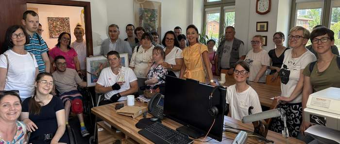 Pokaż zdjęcie: Grupa uśmiechniętych osób, w tym osoby z niepełnosprawnościami, zgromadzona w jasnym biurze z dużym biurkiem na pierwszym planie; na biurku komputer, mikrofon i drukarka.