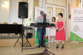 Pokaż zdjęcie: W sali konferencyjnej występ zespołu muzycznego, za kibordem stoi mężczyzna, obok niego dwie kobiety, jedna z nich śpiewa do mikrofonu