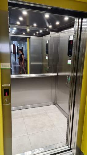 Pokaż zdjęcie: Winda zainstalowana wewnątrz budynku Urzędu Gminy Wągrowiec. Na windzie znajduje się zarówno naklejka informująca, że inwestycja dofinansowana została ze środków PFRON, jak i wyświetlacz z liczbą „1” informującą, że znajduje się ona aktualnie na pierwszym piętrze. W lustrze, wewnątrz windy odbija się postać kobiety idącej korytarzem.