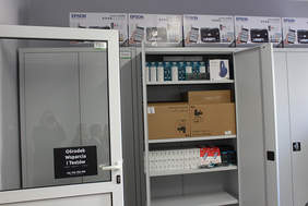 Pokaż zdjęcie: Na zdjęciu 3 szafy, środkowa otwarta, wypełniona sprzętem typu słuchawki, drukarki