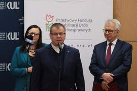 Pokaż zdjęcie: Briefing prasowy od lewej stoi kobieta z ciemnymi włosami w niebieskiej marynarce, przy mikrofonie ksiądz, po prawej starszy mężczyzna w garniturze w okularach