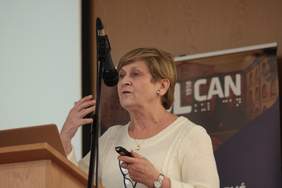 Pokaż zdjęcie: Przy mównicy stoi kobieta. Kobieta w średnim wieku z krótkimi włosami, ubrana w biały sweter, gestykuluje do publiczności