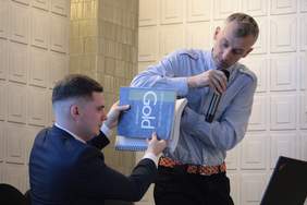 Pokaż zdjęcie: Dwóch mężczyzn trzyma książkę, jeden z nich siedzi, drugi stoi trzymając mikrofon i podtrzymując książkę. Panowie prezentują grubość książki napisanej w języku Braille'a