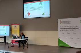 Pokaż zdjęcie: sala konferencyjna po środku stoi biurko przy którym siedzi osoba i przedstawia prezentację dotyczącą centrum informacyjno-doradczym na górze jest wisi duży telebim i wyświetla prezentację 