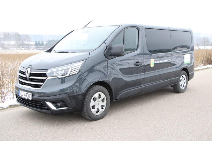 Pokaż zdjęcie: Ciemny mikrobus z logo gminy Iłowo-Osada i PFRON stoi na drodze, w tle zaśnieżone pole