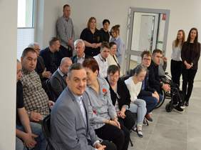 Pokaż zdjęcie: W pomieszczeniu po lewej stronie zdjęcia siedzą na krzesłach osoby. W dalszej części fotografii stoją mieszkańcy i pracownicy Domu Mieszkalnego. 