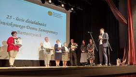 Pokaż zdjęcie: . Na scenie Domu Kultury stoi kilku gości odznaczonych dyplomami, gipsowymi aniołami i kwiatami. Przy mikrofonie zwrócony do dyrektorki Środowiskowego Domu Samopomocy stoi dyrektor PFRON.   