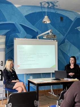 Pokaż zdjęcie: Dwie kobiety siedzą przy stole, na którym znajduje się laptop, za nimi tablica z wyświetloną prezentacją.