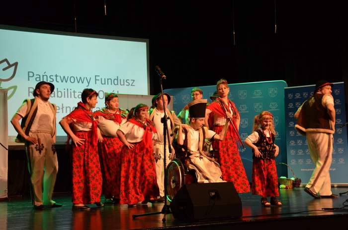 Pokaż zdjęcie: W pomieszczeniu na scenie stoi 12 osób w strojach góralskich w tym jedna jest na wózku inwalidzkim, w tle na dużym ekranie logo PFRON