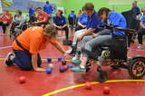 Uczestnicy na wózkach inwalidzkich w hali w trakcie gry w boccię, zawowdnicy są w niebieskich koszulkach, obok sędzia w pomarańczowej koszulce mierzy odległości