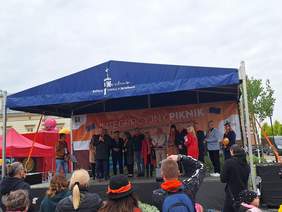 Scena na rynku, zadaszona, na niej grupa ludzi, jedna osoba przemawia, pod scena stoją zebrani