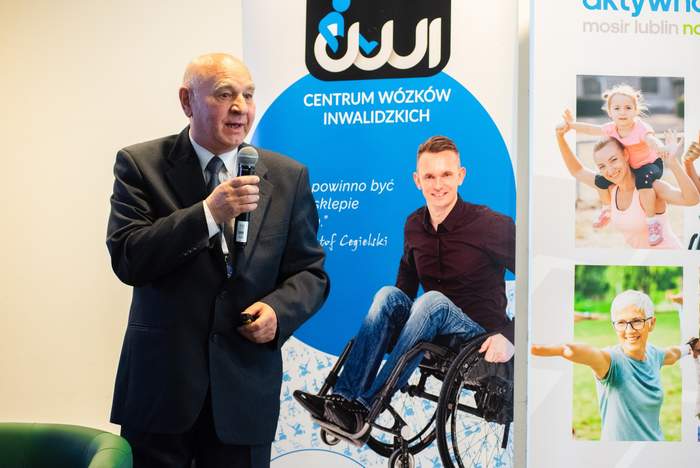 Pokaż zdjęcie: Przy mikrofonie stoi starszy mężczyzna w garniturze. W tle rollup z osobą na wózku.