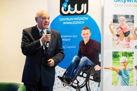 Pokaż zdjęcie: Przy mikrofonie stoi starszy mężczyzna w garniturze. W tle rollup z osobą na wózku.