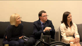 Pokaż zdjęcie: Na kanapach siedzą dwie kobiety i mężczyzna. Rozmawiają ze sobą. Przed nimi stoi wózek inwalidzki. 