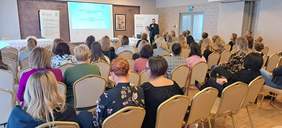 Sala konferencyjna, zapełniona osobami siedzacymi na krzesłach - tyłem do obiektywu. Słuchają prelegenta, który omawia prezentację, wyświetlaną na ekranie.