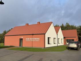 Pokaż zdjęcie: Nowoczesny budynek z czerwonym dachem i elewacją w kolorze ceglastym oraz jasnym. Na ścianie napis „Dom Rain Mana – Rodzinny Ośrodek Pomocy”. Obiekt stoi na zielonym terenie, obok podjazd i zaparkowany samochód.
