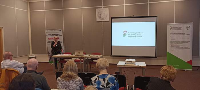 Pokaż zdjęcie: Samodzielny Referent ds. CIDON Anna Przybylska a w trakcie wystąpienia, na tle prezentacji z logo PFRON, z mikrofonem w ręku, widać salę wypełnioną goścmi