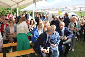 Pokaż zdjęcie: Ciepły słoneczny dzień, pod namiotem na ławkach siedzą uczestnicy pikniku dnia rodzin