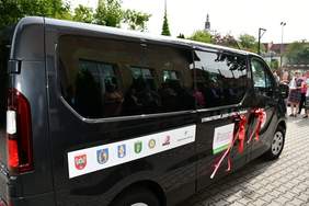 Pokaż zdjęcie: Czarny mikrobus z naklejką informującą, że pojazd przystosowany jest do przewozu osób z niepełnosprawnością. Na samochodzie znajdują się także logotypy instytucji, które dofinansowały zakup busa oraz nazwa Stowarzyszenia „Razem Łatwiej” w Wolsztynie.