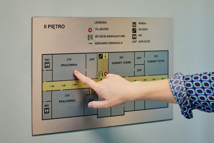 Pokaż zdjęcie: Na górze napis „II piętro” wraz z legendą opisującą poszczególne symbole znajdujące się na planie. Poniżej kobiecy palec, który dotyka oznaczenie w alfabecie Braille’a informujące, że w tym miejscu na planie znajduje się pracownia o numerze 214.