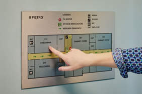 Pokaż zdjęcie: Na górze napis „II piętro” wraz z legendą opisującą poszczególne symbole znajdujące się na planie. Poniżej kobiecy palec, który dotyka oznaczenie w alfabecie Braille’a informujące, że w tym miejscu na planie znajduje się pracownia o numerze 214.