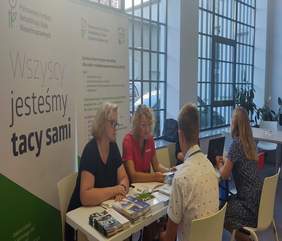 Pokaż zdjęcie: sala obsługi klientów na zdjęciu stoisko PFRON dwie pracownice udzielają informacji klientom. Z tyłu logo PFRON