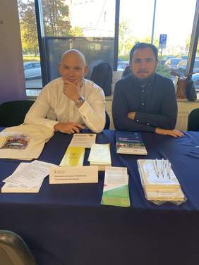 Pokaż zdjęcie: Dwóch mężczyzn siedzi przy stole, na którym rozłożone są ulotki, broszury informatory, torby i długopisy. Ubrani są w koszule. Znajdują się w pomieszczeniu. Za nimi okno, na zewnątrz parking. 