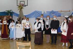 Pokaż zdjęcie: 8 kobiet i 6 mężczyzn stoją na scenie. Niektórzy trzymają dyplomy s serduszkiem. Za nimi gwiazda betlejemska.