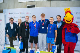 Pokaż zdjęcie: Zdjęcie grupowe po ceremonii medalowej zawodów pływackich. Na tle z logo PZSN Start, Ministerstwa Sportu i Turystyki oraz PFRON, stoi siedem osób: dwóch mężczyzn, kobieta, maskotka i trzech młodych mężczyzn. Maskotka, duża, czerwono-niebiesko-żółta papuga w koronie, stoi po lewej stronie, opierając rękę na ramieniu młodego mężczyzny na wózku inwalidzkim, który stoi na miejscu z numerem 2 (drugie miejsce). Pośrodku, na miejscu 1 (pierwsze miejsce), siedzi na podłodze uśmiechnięty, rudowłosy młody mężczyzna. Po prawej, na miejscu 3 (trzecie miejsce), klęczy kolejny młody mężczyzna. Cała trójka ma na szyjach medale na wstążkach. Obok maskotki i za zawodnikami stoi kobieta w białej koszulce. Po lewej i prawej stronie grupy stoją dwaj starsi mężczyźni w strojach sportowych.