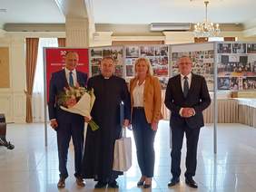 Pokaż zdjęcie: Zdjęcie kobiety, księdza i dwóch mężczyzn w garniturach. Ksiądz trzyma bukiet kwiatów i siatki z prezentami