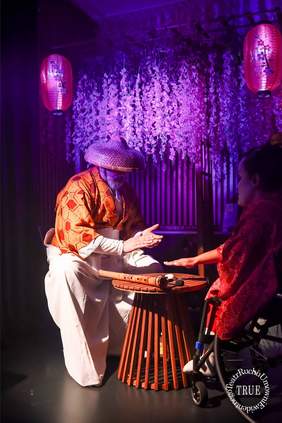 Pokaż zdjęcie: Pionowe ujęcie w fioletowym oświetleniu, przedstawiające scenę z dwoma aktorami. Mężczyzna po lewej stronie, ubrany w luźny, pomarańczowo-biały strój w geometryczne wzory i duży, stożkowy kapelusz, siedzi na małym, ażurowym, drewnianym stoliku. Trzyma w dłoniach mały instrument lub rurkę bambusową. Po prawej, na wózku inwalidzkim, siedzi aktorka w czerwonym kimonie, wyciągająca rękę w kierunku dłoni mężczyzny. W tle wiszą girlandy z fioletowych kwiatów i dwie różowe, papierowe lampiony.