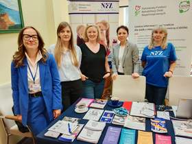 Pięć kobiet stoi na tle banerów: NFZ i PFRON. Przed nimi jest stół, na którym leżą różne ulotki, poradniki, broszury i inne materiały dotyczące zagadnień związanych ze zdrowiem. 