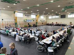 Pokaż zdjęcie: Na sali gimnastycznej rozłożone jest po obrzeżach kilka długich stołów w krztałt litery U. Siedzą przy nich osoby jedzą ciasto. Na środku sali stoi dwóch mężczyzn w garniturach.