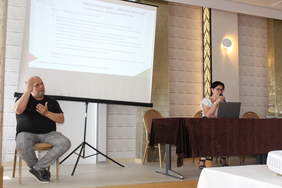 Pokaż zdjęcie: W sali konferencyjnym po lewej stronie siedzi na krześle tłumacz języka migowego, po prawej za stołem prezydialnym siedzi kobieta, druga stoi z mikrofonem, w tle po lewej na ekranie wyświetlana jest prezentacja