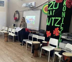 Pokaż zdjęcie: Kobieta z mikrofonem trzyma teczkę z listem gratulacyjnym. Na sali są także krzesła oraz bębny. W tle znajduje się ekran, na którym wyświetlono prezentację. Po prawej stronie ustawiona jest ścianka z kwiatami oraz napisem: „20 lat WTZ Koniczynka”. 