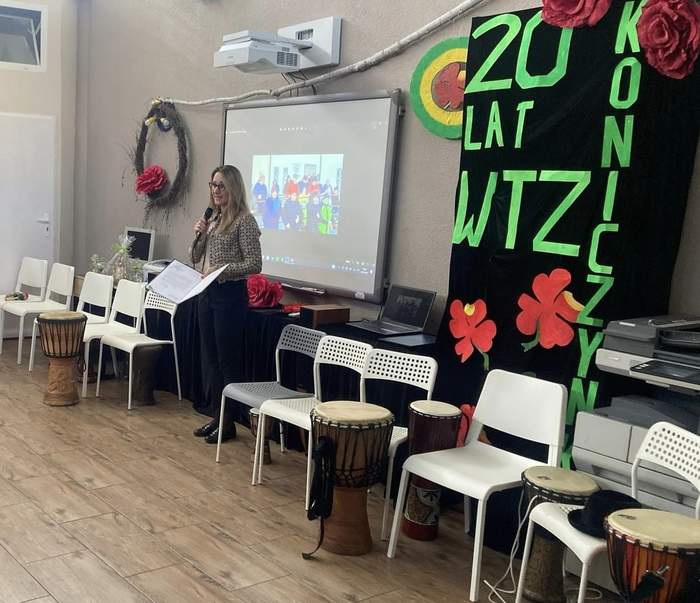 Pokaż zdjęcie: Kobieta z mikrofonem trzyma teczkę z listem gratulacyjnym. Na sali są także krzesła oraz bębny. W tle znajduje się ekran, na którym wyświetlono prezentację. Po prawej stronie ustawiona jest ścianka z kwiatami oraz napisem: „20 lat WTZ Koniczynka”. 