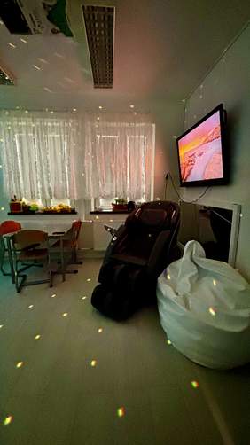 Pokaż zdjęcie: Pomieszczenie relaksacyjne w ośrodku wyposażone w fotel masujący, pufę typu worek, telewizor na ścianie oraz stolik z krzesłami; widoczne są elementy oświetlenia dekoracyjnego.