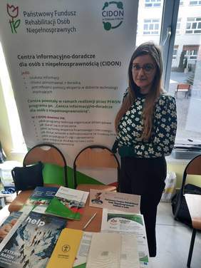 Pokaż zdjęcie: W sali wystawienniczej znajduje sie stoisko Państwowego Funduszu Rehabilitacji Osób Niepełnosprawnych. Obok stoiska stoi młoda kobieta - pracownica PFRON. Na stole leżą różne informatory, poradniki, ulotki i broszury. 