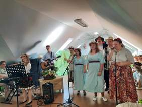 Pokaż zdjęcie: Na scenie artyści wykonują koncert. Niektóre osoby grają na instrumentach muzycznych, a inne śpiewają do mikrofonów. Kobiety są ubrane w kolorowe sukienki, a mężczyźni w koszule. 