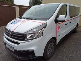 Pokaż zdjęcie: Zbliżenie białego busa na przedniej masce naklejony duży niebieski znaczek osoby na wózku inwalidzkim i logo Caritas samochód stoi na tle garaży i budynku