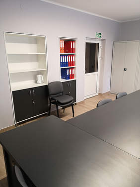 Pokaż zdjęcie: Duży jasny pokój, na środku stoją dwa czarne stoły połączone w kształt litery T, przy ścianie stoją dwa białe regały na segregatory, jeden jest pusty, drugi wypełniony segregatorami